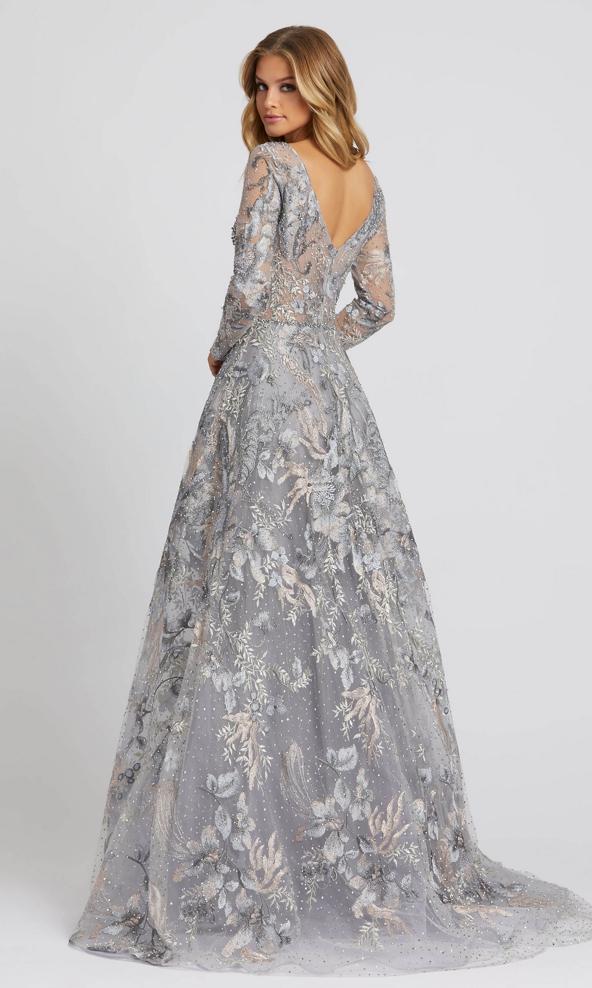 A woman models the Long Sleeve Long Formal Dress: Mac Duggal 20214—a floor-length, silver-gray gown with floral patterns, long sleeves, and a low-cut back—against a plain background.